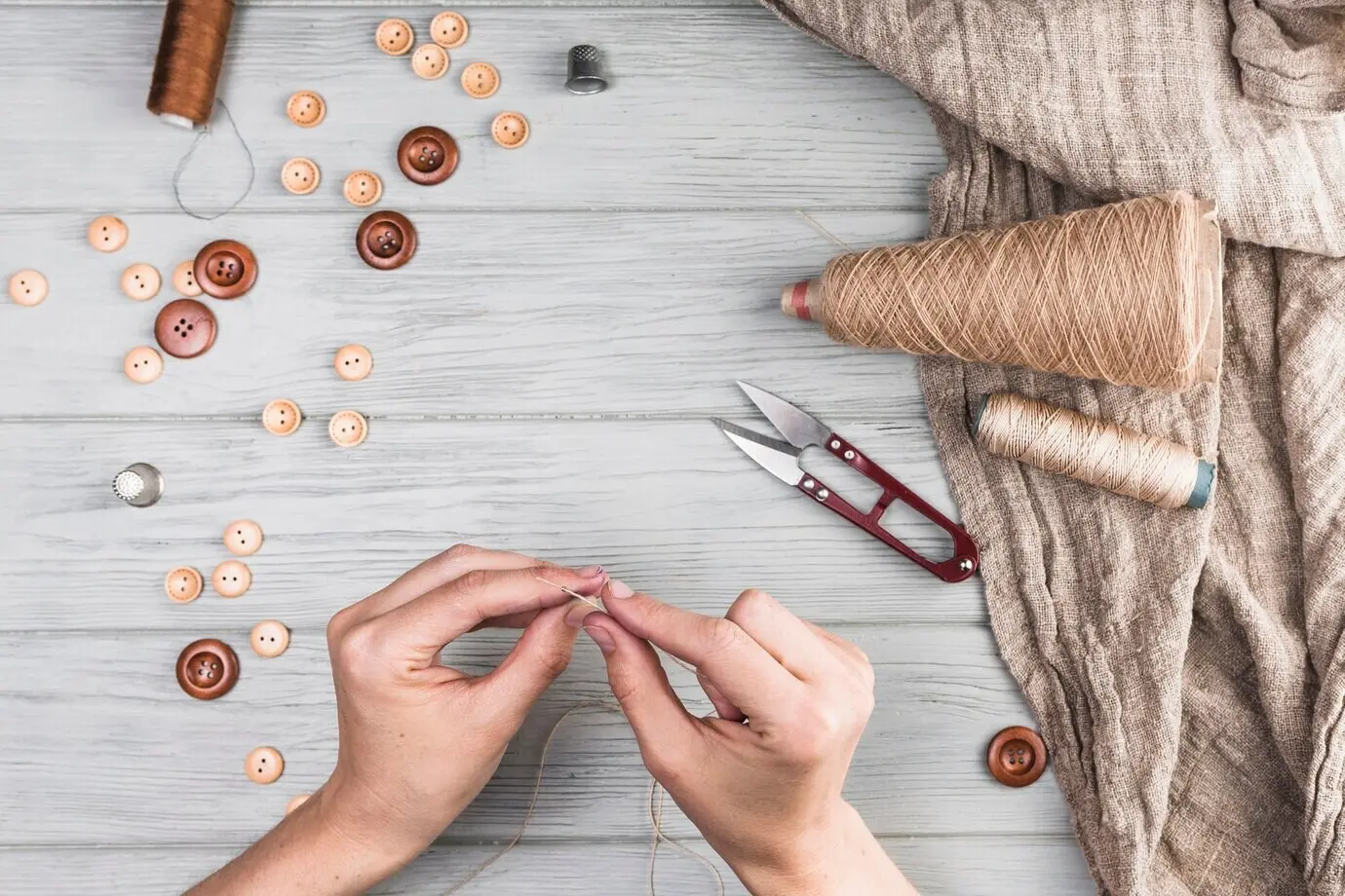 Nahaufnahme der Hand einer Frau, die einen Faden in eine Nadel einfädelt; mit Knopf, Cutter und Stoff auf hölzernem Untergrund.